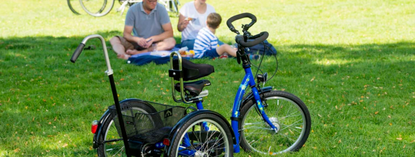 Dreirad von Schuchmann im Vordergrund. Im Hintergrund picknickt eine Familie mit Kind mit Behinderung.